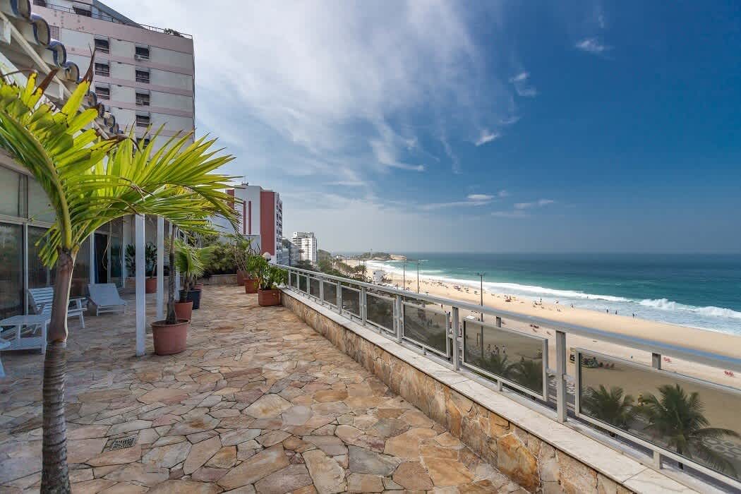 The open terrace of the Ipanema penthouse, paved in irregular stone with potted palms and white loungers, looking east along the beach towards the rocks of Arpoador