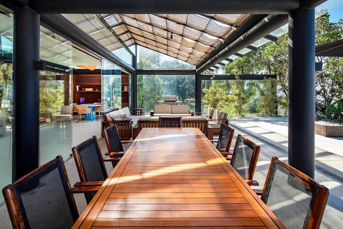 A long slatted wooden dining table with dark mesh-backed chairs under the São Conrado villa's glass and fabric-shaded roof, with outdoor sofas and the garden beyond