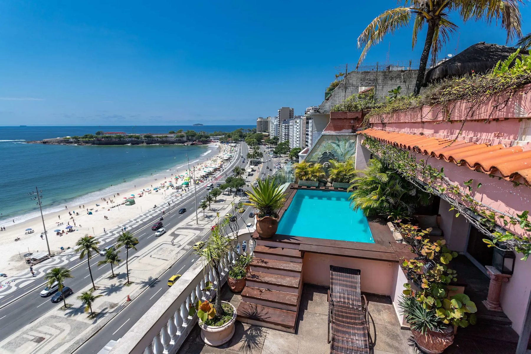 A rooftop terrace with a small turquoise pool on a wooden deck, potted palms, a lounger and a stone balustrade, high above a beachfront avenue, white sand and a headland.