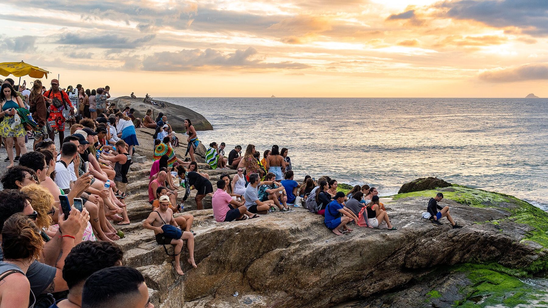 A crowd of people sitting and standing on a large sloping rock by the sea, many holding up phones, facing an orange and grey sunset sky over the ocean.