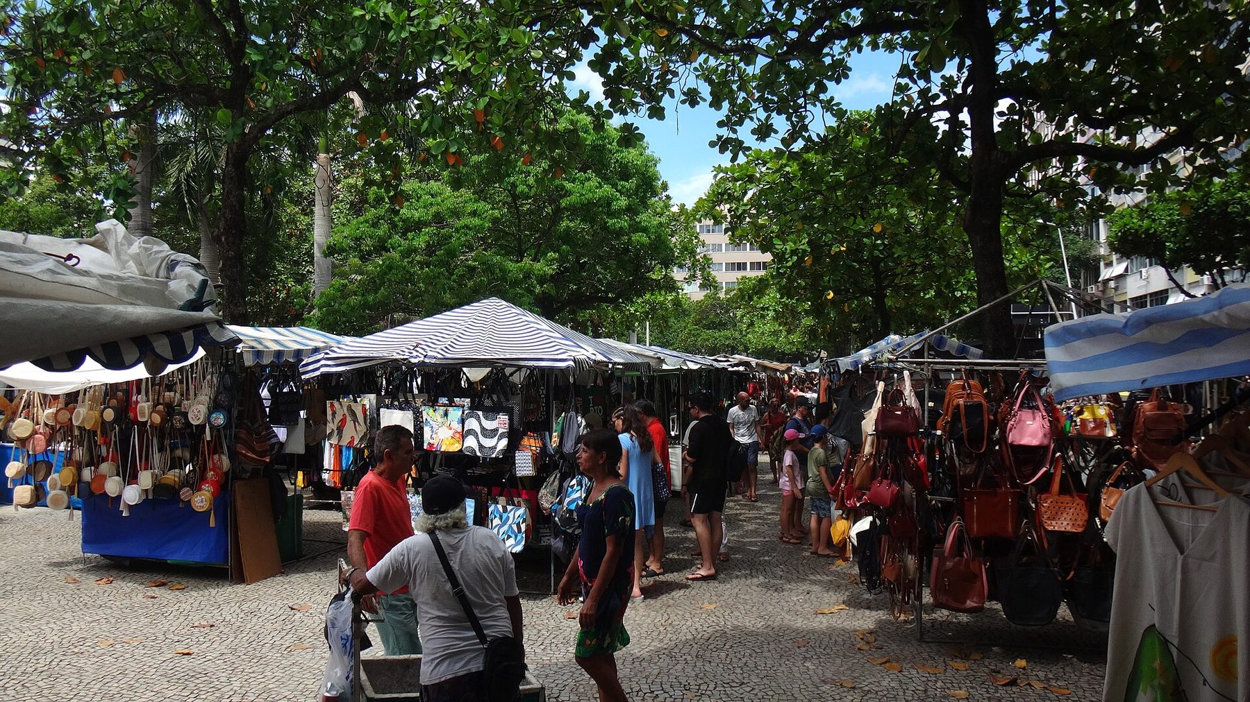 An open-air market under large trees, with striped canopies, stalls hung with leather bags, baskets and printed fabrics, and shoppers walking on a stone-paved square.