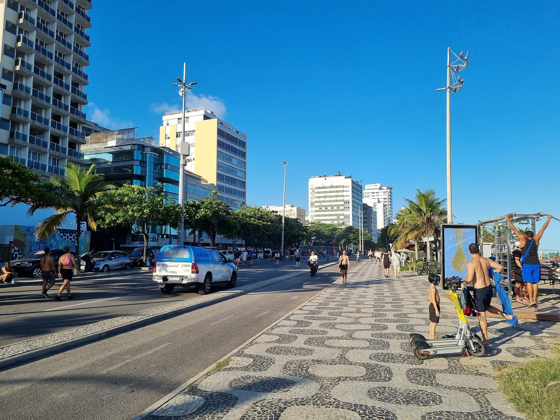 A beachfront avenue with a patterned black and white stone pavement, walkers, a man using an outdoor exercise frame, a police pickup truck, palms and apartment buildings under a clear blue sky.