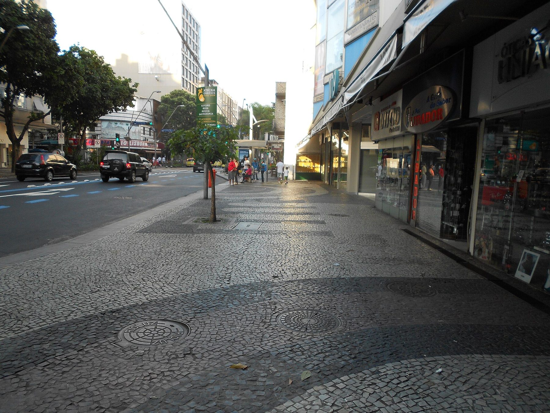 A wide city pavement of small grey and white stones beside shopfronts with signs, a young tree, a street with cars and a traffic light, and a few pedestrians in the distance.