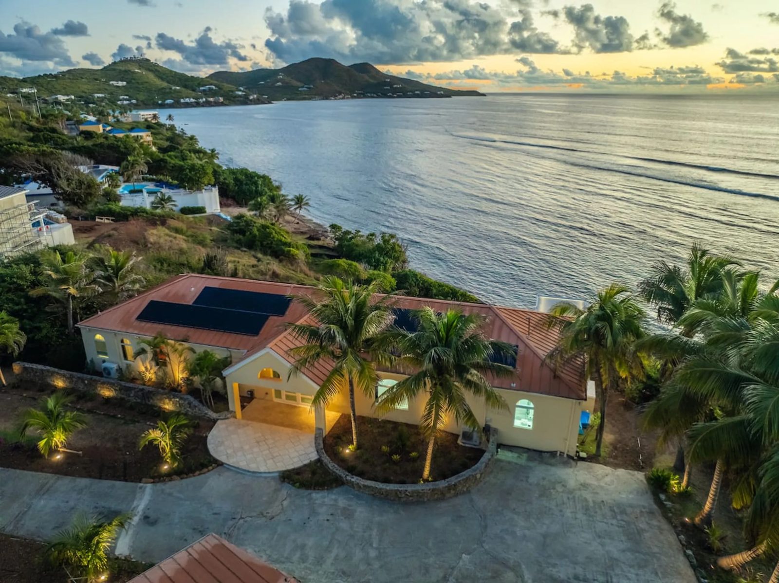 The Cerulean on St. Croix from the land side: a pale yellow villa with a copper roof and solar panels, palms and a circular drive, its garden lights on, and the coast and hills beyond