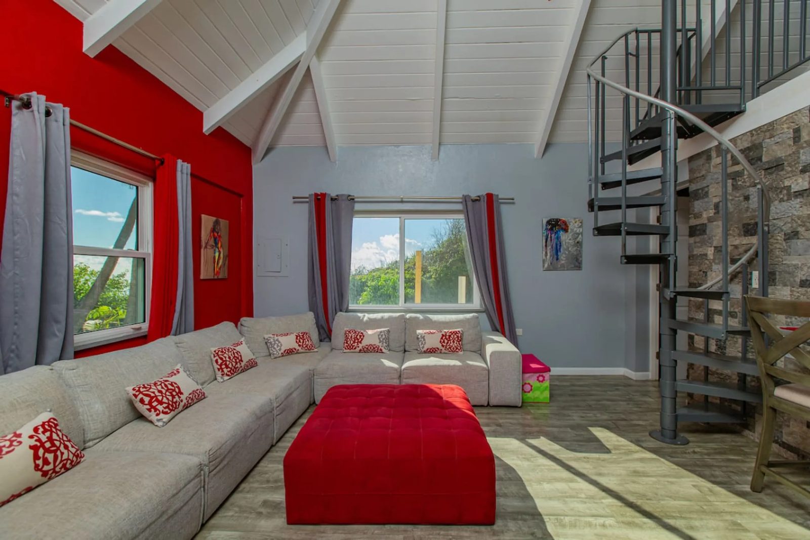 The living room of The Cerulean's guest cottage, with a large white sectional sofa, a red ottoman, a red accent wall and a spiral stair to a loft