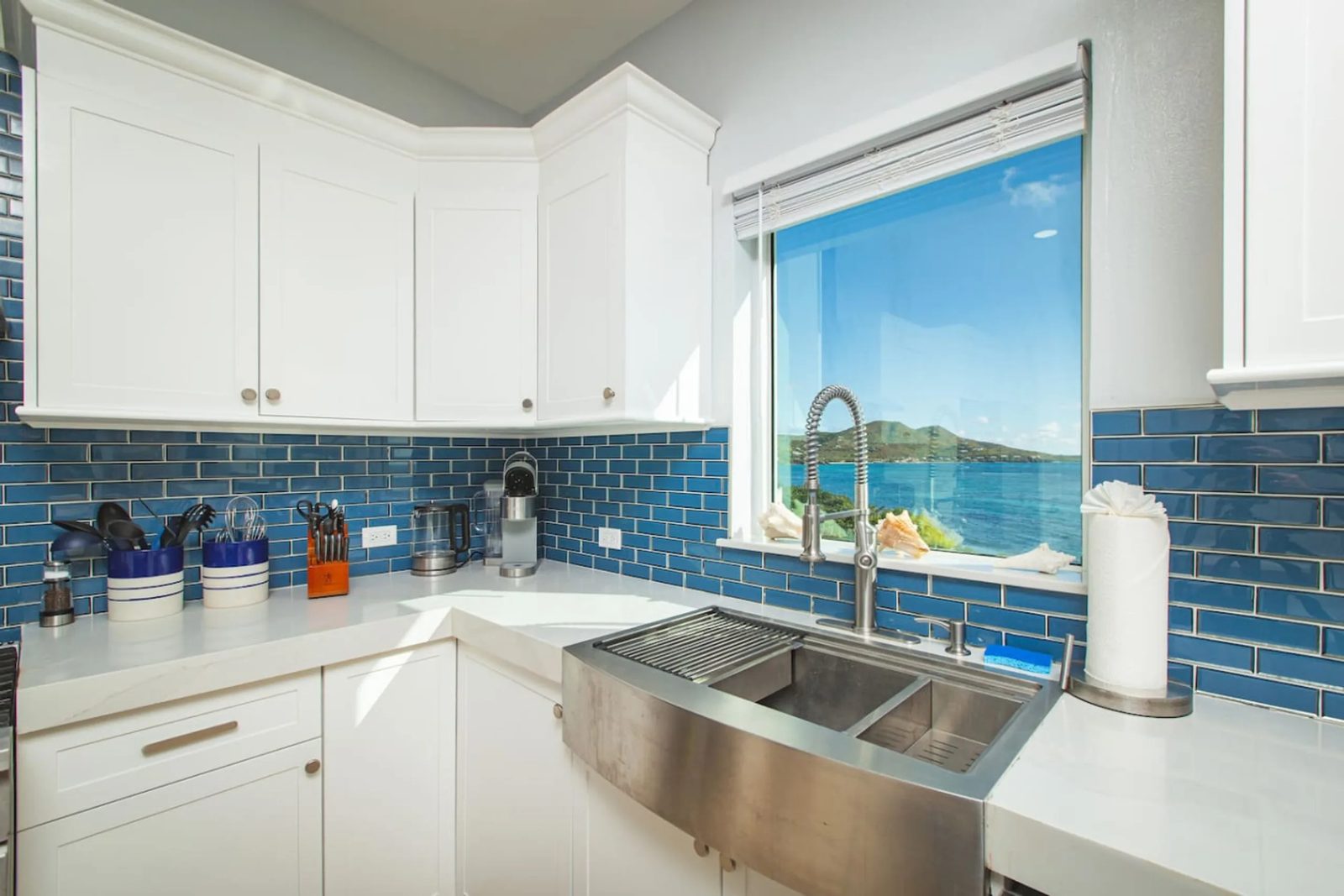 The kitchen sink at The Cerulean villa under a window looking out over the water, with blue tiles and white cabinets