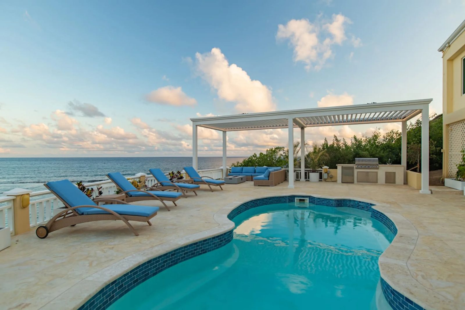 The free-form pool at The Cerulean by day, with blue loungers on the deck, a white louvred pergola and the sea beyond
