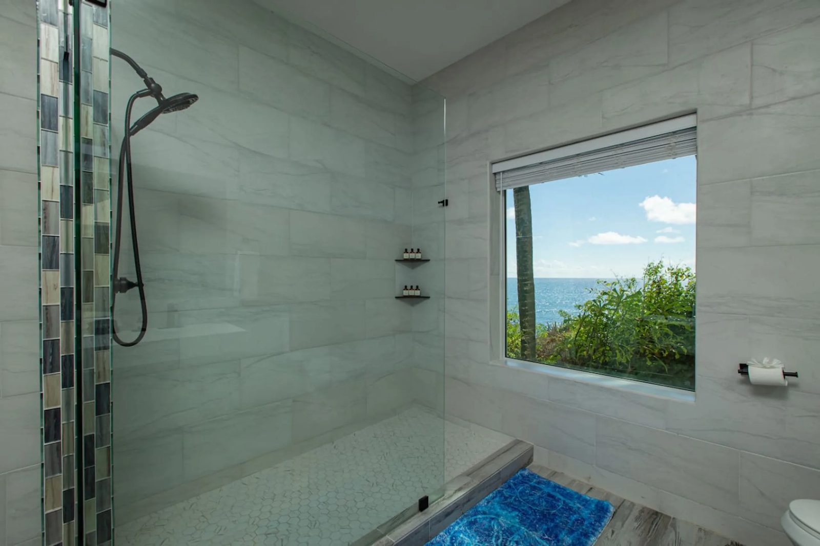 A tiled walk-in shower at The Cerulean with a window looking out over green planting to the sea