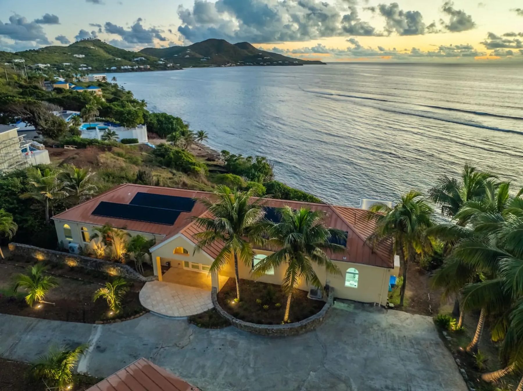 Aerial view of a single-storey wing with a copper-coloured roof and solar panels, a circular paved drive and palm trees, with the coast, neighbouring houses and green hills curving away under a cloudy evening sky.
