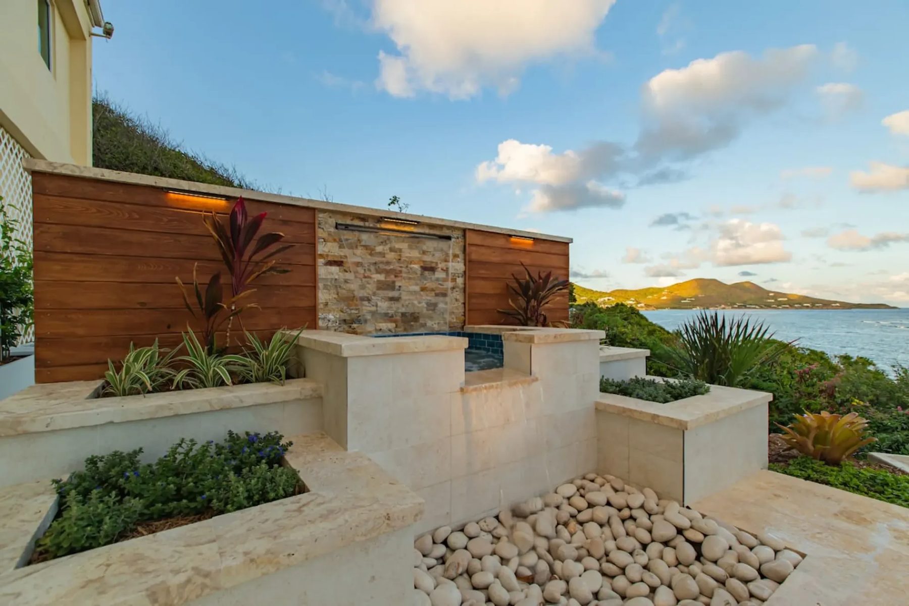 A built garden water feature: a thin sheet of water falls from a stacked-stone panel set in a timber wall into a small tiled basin and a bed of white pebbles, with planters and the sea behind.