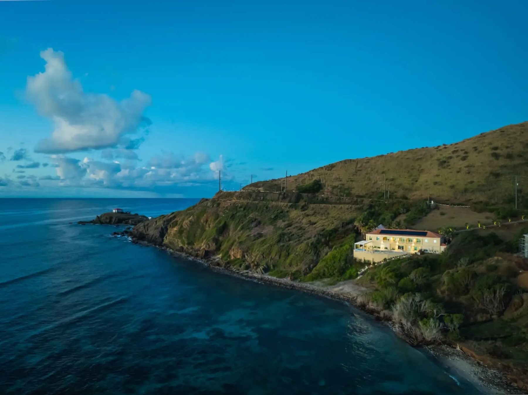 Aerial view of a cream two-storey villa on a green hillside above a narrow stony shore, with deep blue water, a rocky point and a small islet beyond under a blue sky.