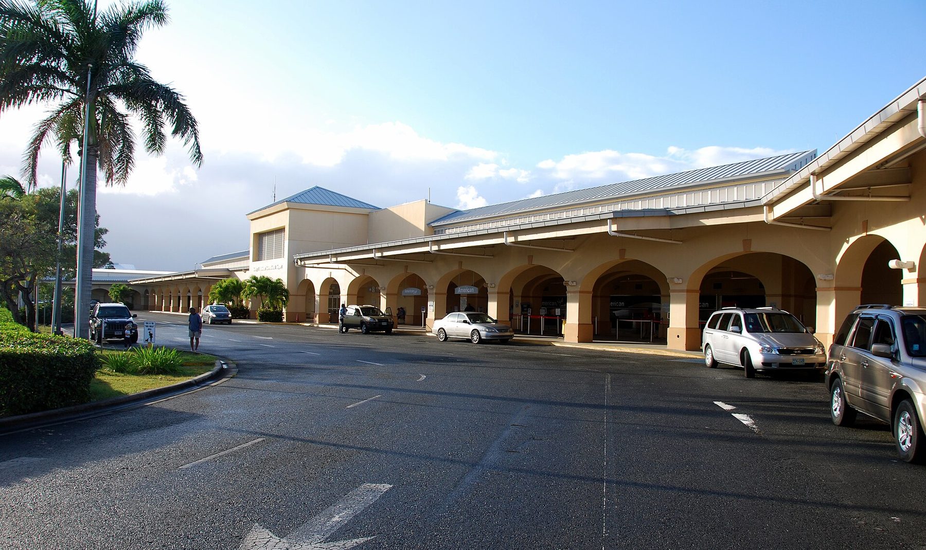 The kerbside front of a cream airport terminal with a long arcade of arches, a metal roof and airline signs, cars parked along the road and a palm tree at left; a few small figures stand in the distance.