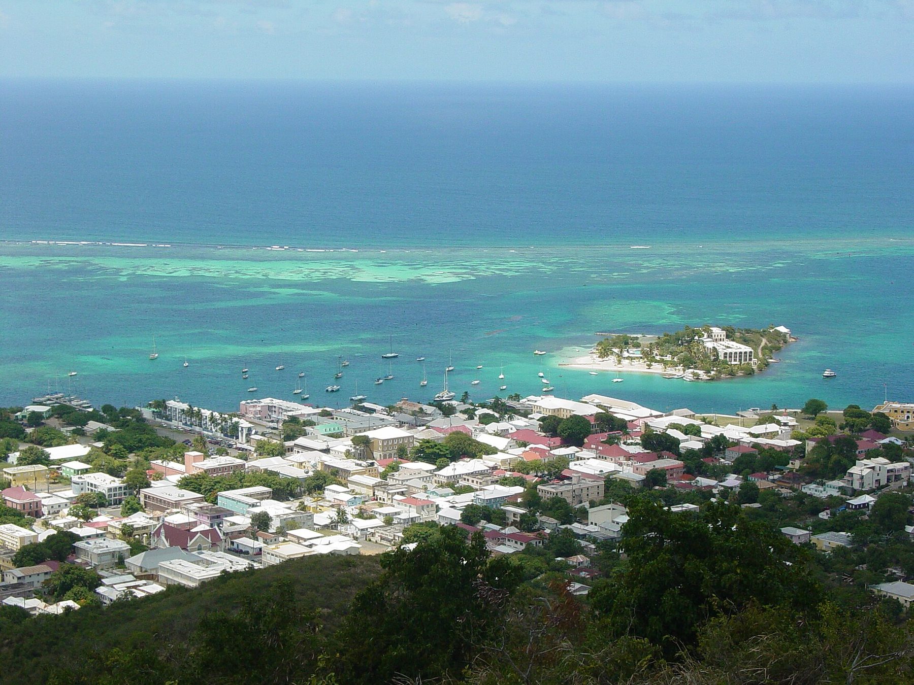 View from a hillside over the rooftops of Christiansted to its harbour, with moored sailboats, a small wooded cay with a white hotel building and beach, pale green shallows, a line of reef and open sea.