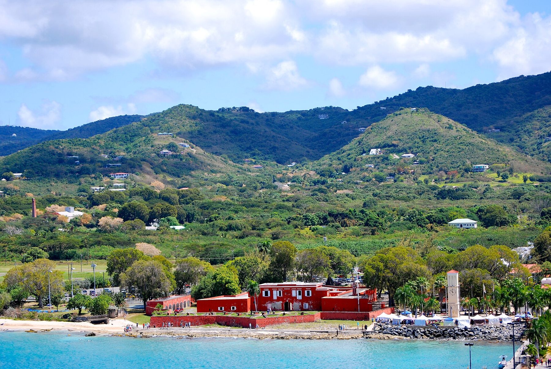 A red-walled fort and a white clock tower on a shoreline of pale water and rocks, with trees, market tents and scattered houses on green hills behind under a cloudy sky.