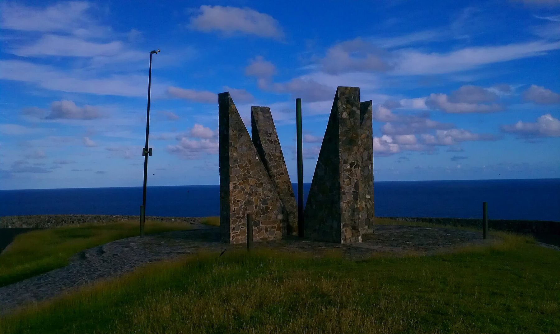 A stone monument of tall angled masonry slabs standing on a grassy headland, with a lamp post beside it and a dark blue sea and cloud-streaked sky behind; the picture is dark and strongly saturated.