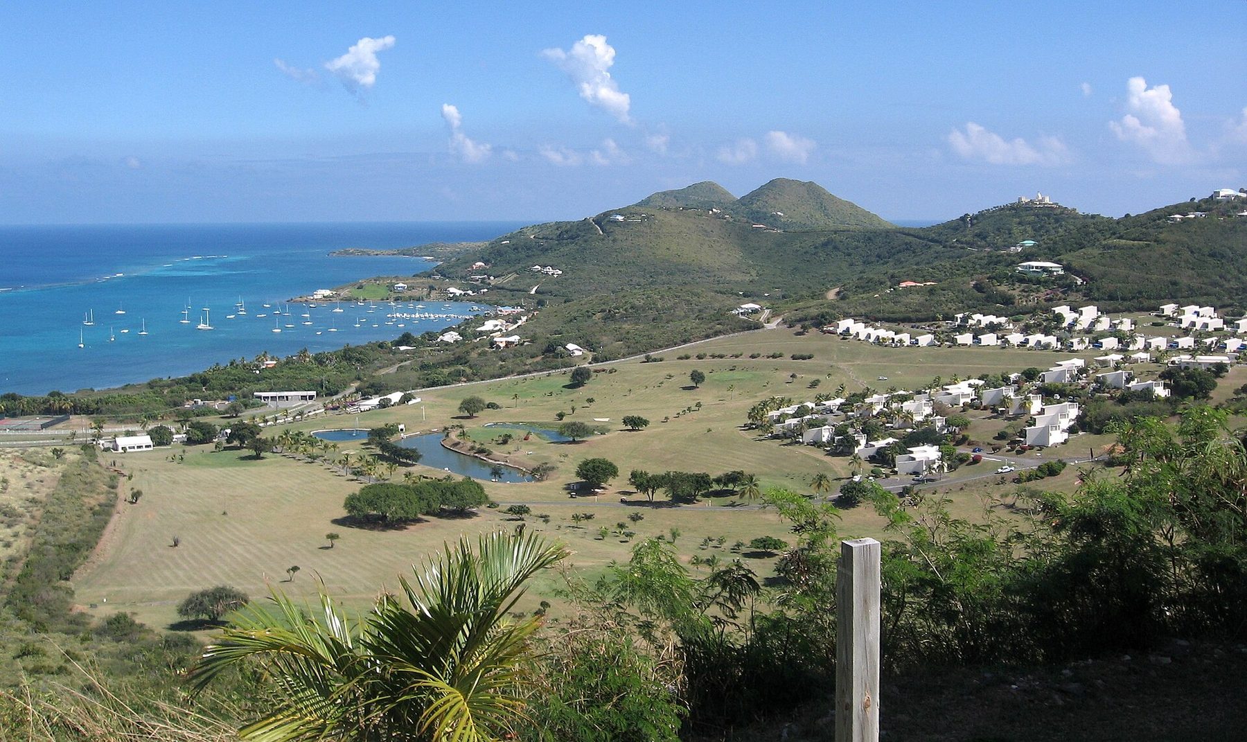 View from a hill over a golf course with ponds and white low-rise buildings to a bay of turquoise water with many moored sailboats, and rounded green hills along the coast beyond.