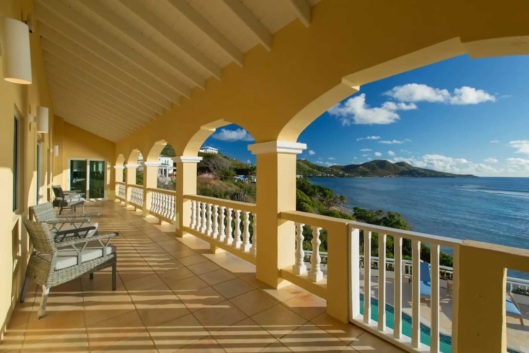 A long covered upper veranda with terracotta floor tiles, a white beamed ceiling, cream arches and woven chairs, looking along the coast to green hills across blue water in sunshine.