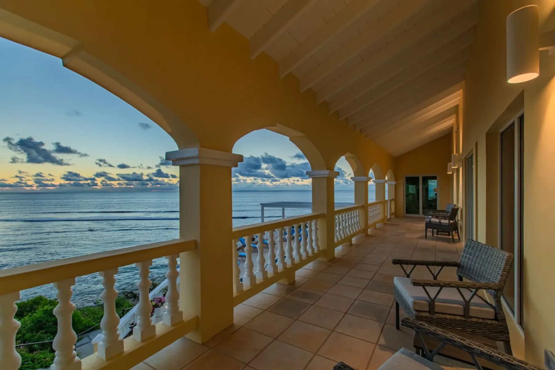 A covered upper veranda in dim early light, with cream arches, a white balustrade, woven chairs and wall lamps, and a pale orange glow along the horizon over a grey-blue sea.