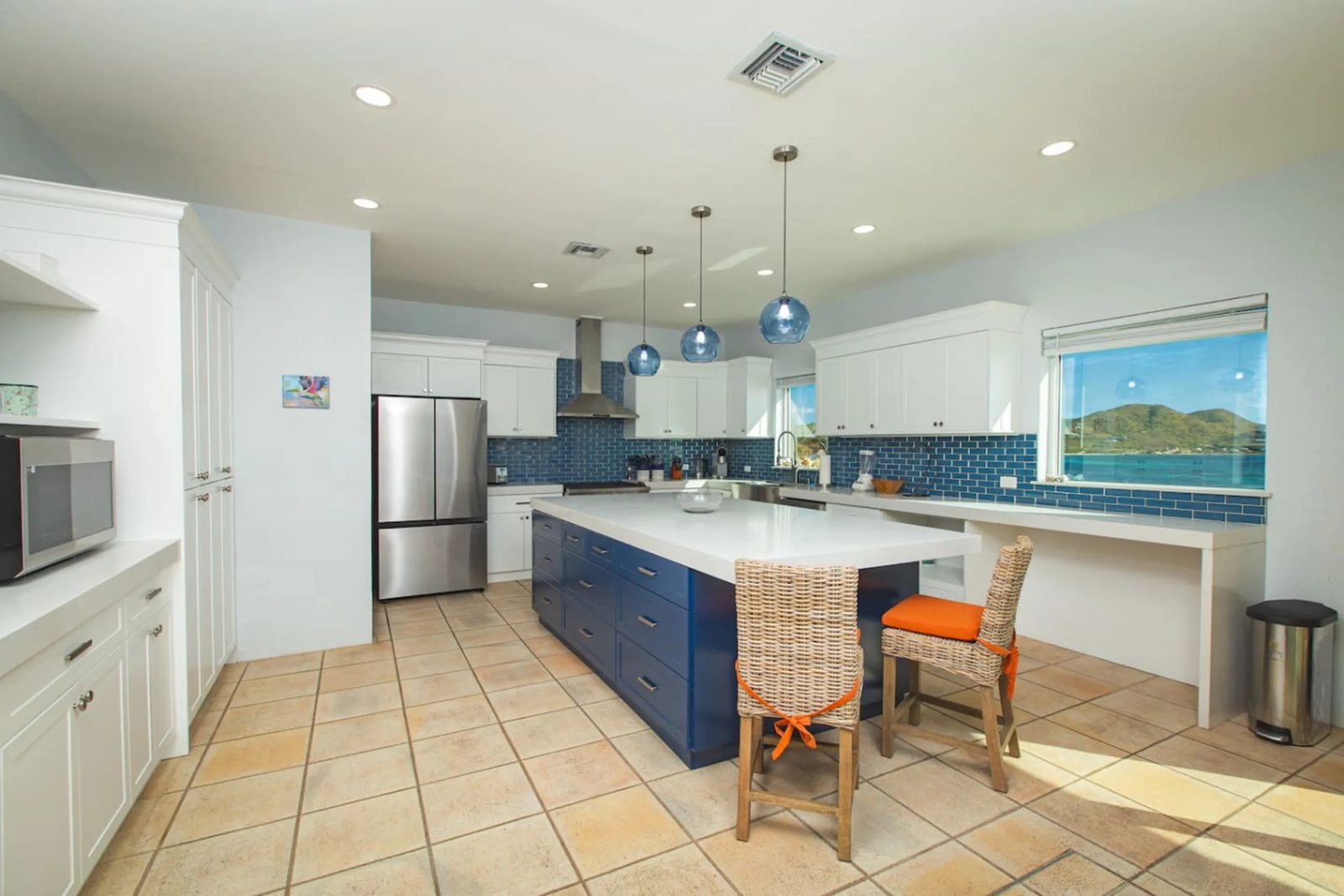 The kitchen at The Cerulean on St. Croix: white cabinets and blue tiles, a long blue island with two woven stools, a steel refrigerator and range under a steel hood, and a window onto the bay and a green headland