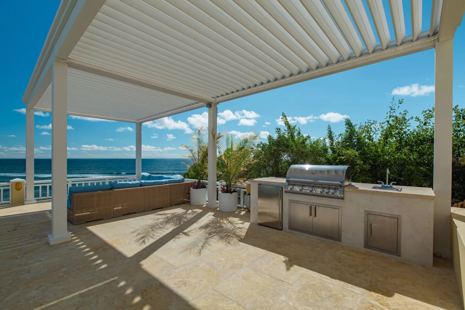 The outdoor kitchen at The Cerulean under a white louvred pergola: a built-in steel grill, a small steel fridge and a sink set into a stone counter, a cushioned lounge, and the sea beyond the balustrade