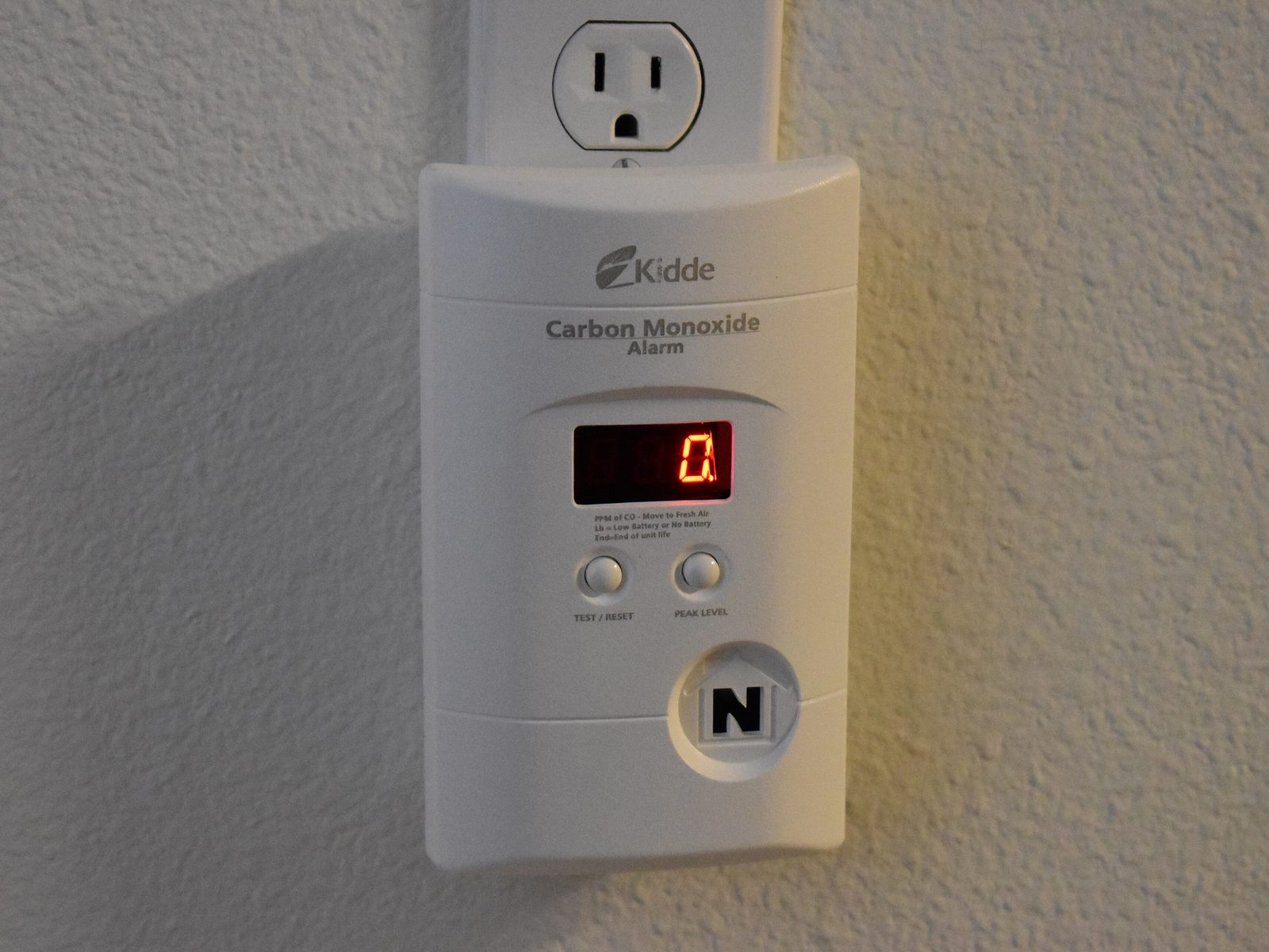 A plug-in carbon monoxide alarm below a wall socket, its red display reading zero, with test and peak-level buttons