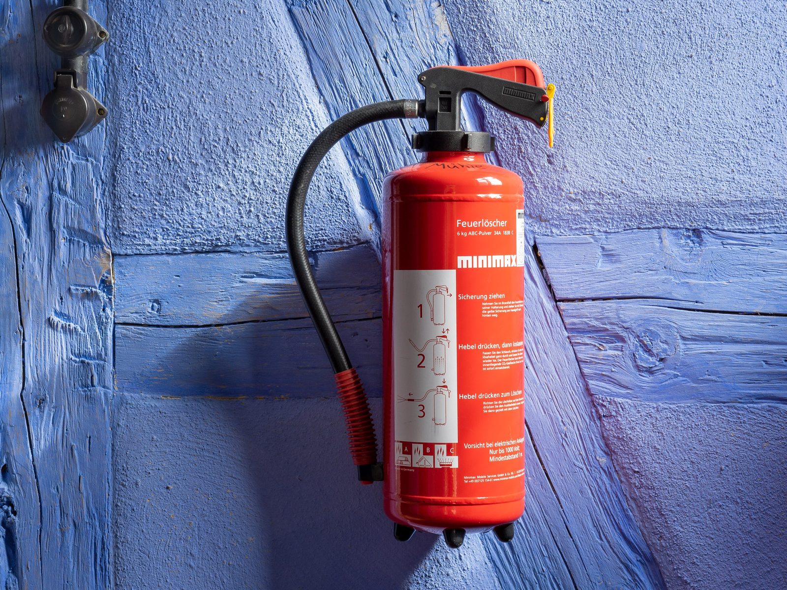 A red multipurpose powder fire extinguisher with a black hose and three numbered pictogram instructions, hanging on a bracket against a blue-painted timber and plaster wall