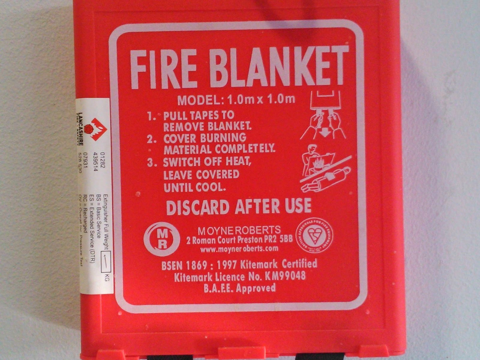 A red wall-mounted fire blanket case printed with instructions: pull tapes to remove blanket, cover burning material completely, switch off heat, leave covered until cool, discard after use