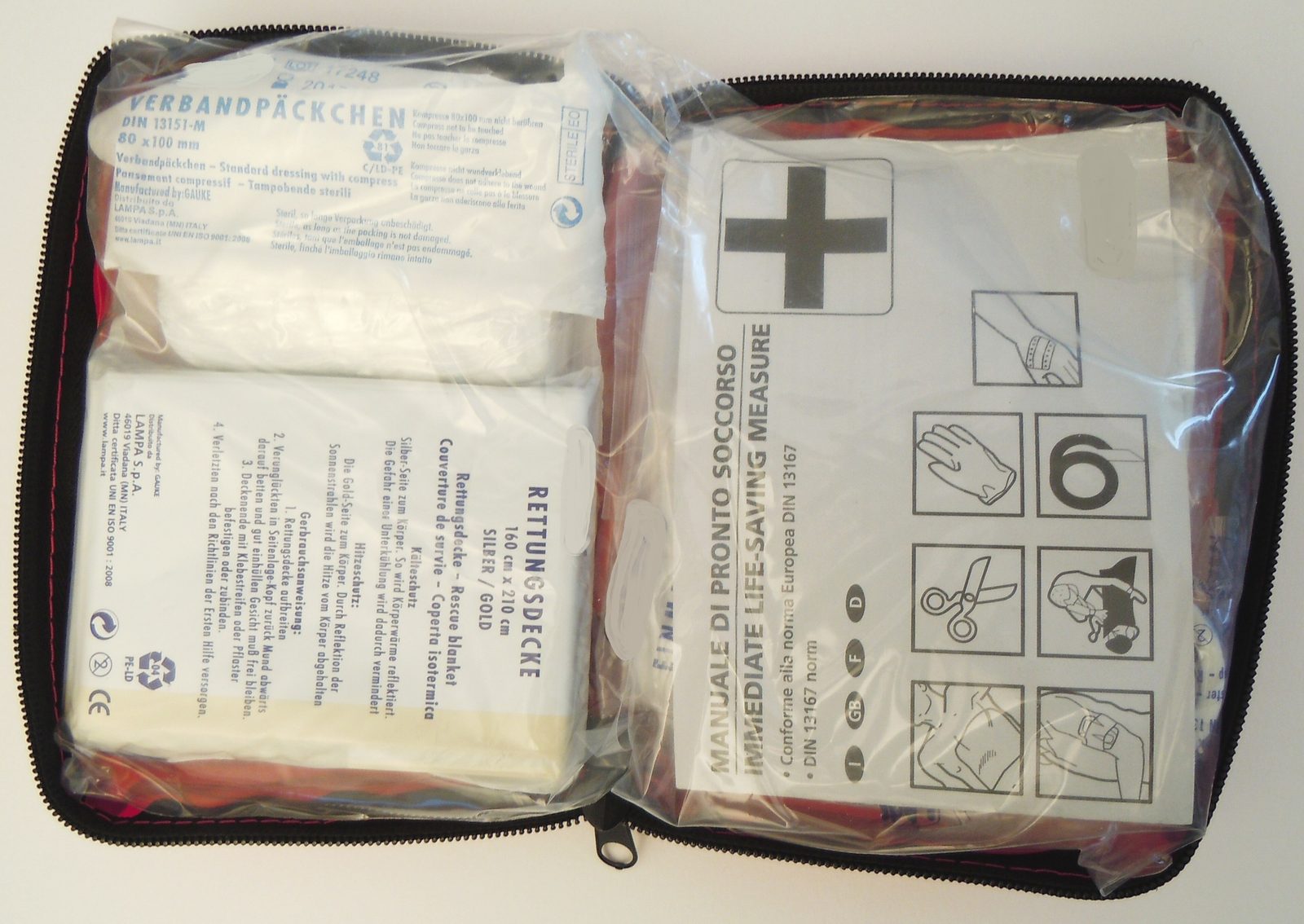 An open first-aid kit in a zipped case, holding a packed compress dressing, a folded foil rescue blanket and an illustrated booklet of first-aid steps