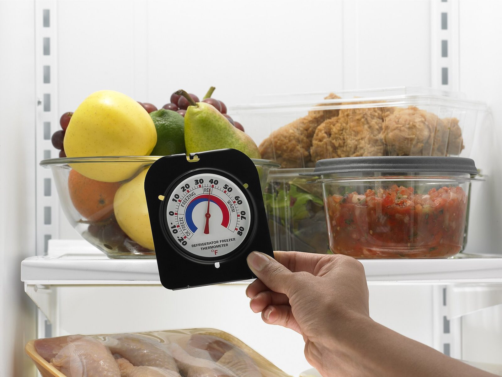A hand holding a dial refrigerator and freezer thermometer marked in Fahrenheit in front of a fridge shelf with a bowl of fruit, lidded containers and raw chicken on the shelf below