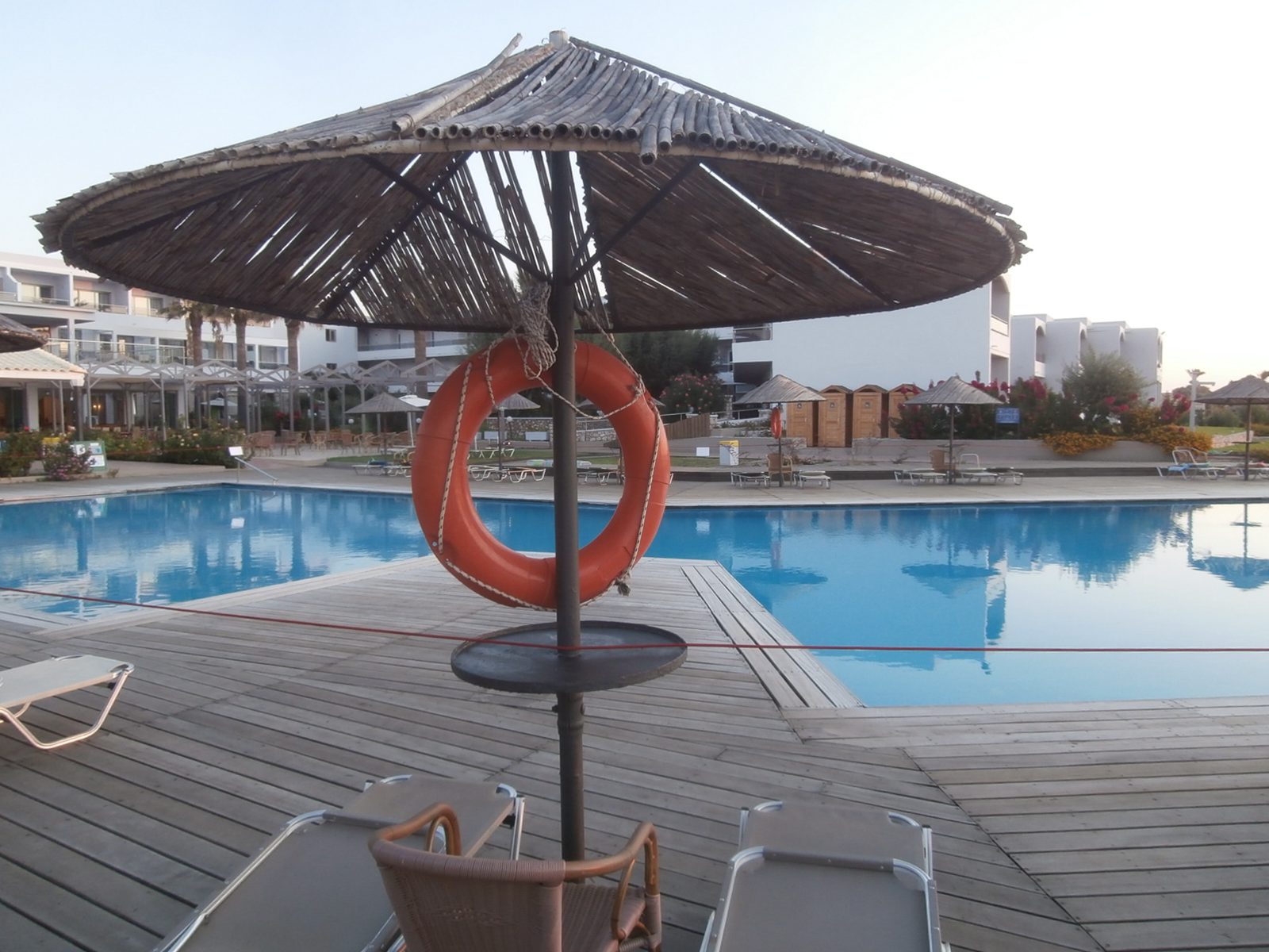 An orange life ring hanging from a thatched parasol beside a hotel swimming pool on Rhodes, with loungers on the wooden deck and white buildings beyond