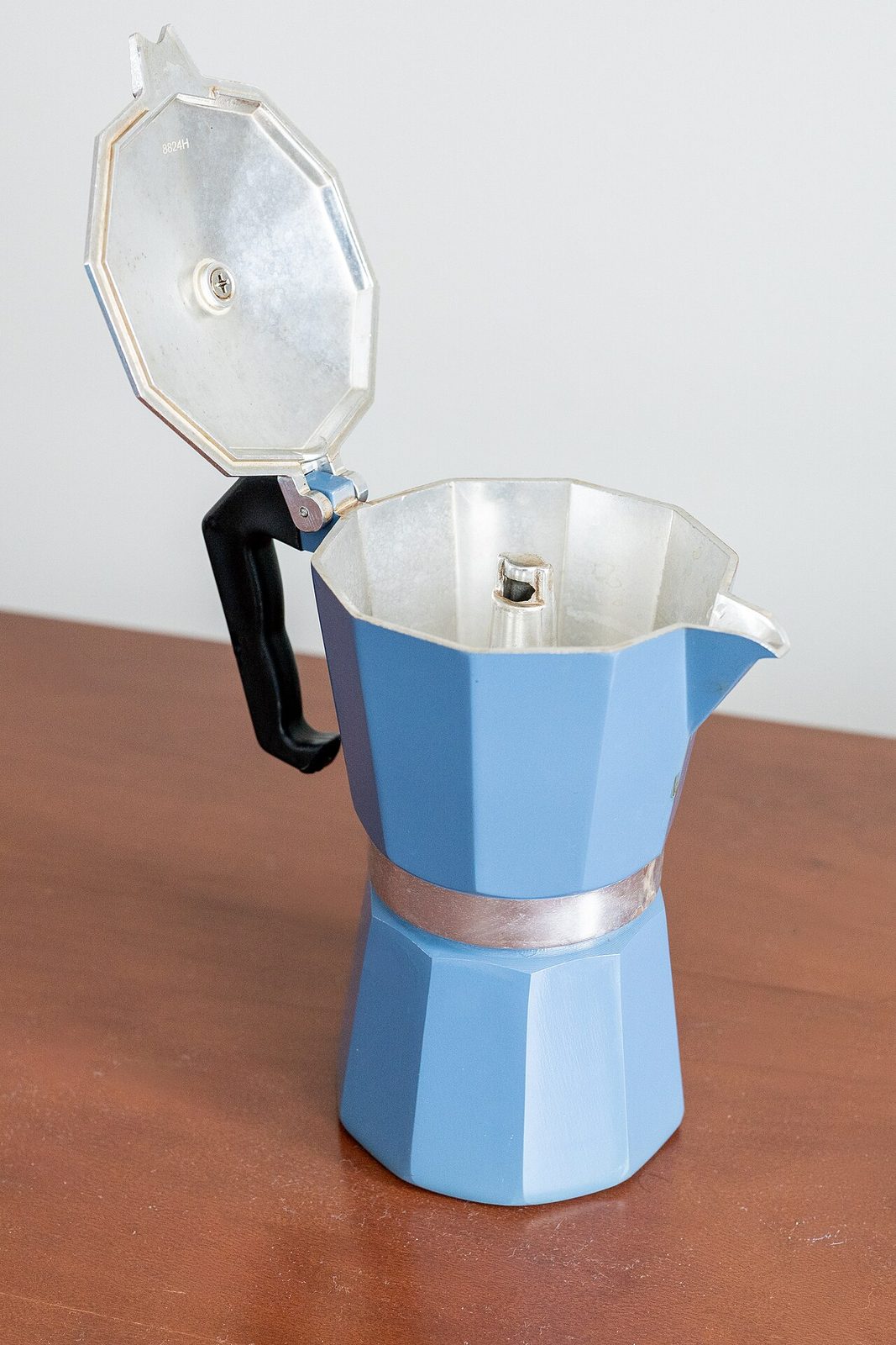 A blue aluminium stovetop moka pot with a black handle and its hinged lid open, standing on a wooden table against a white wall