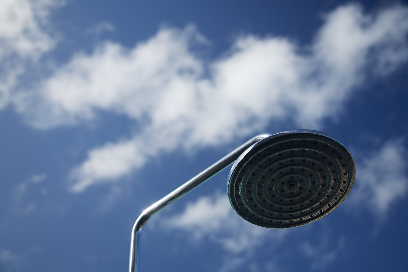 A round chrome rain shower head with rings of small nozzles on a bent chrome arm, seen from below against a blue sky with white clouds