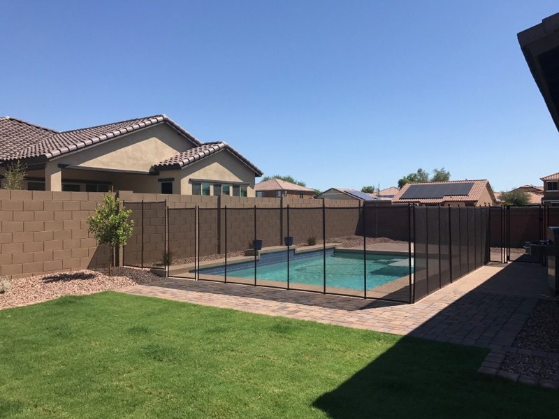 A removable black mesh safety fence around a backyard swimming pool in Arizona, standing between the pool and the lawn, with block walls and houses beyond