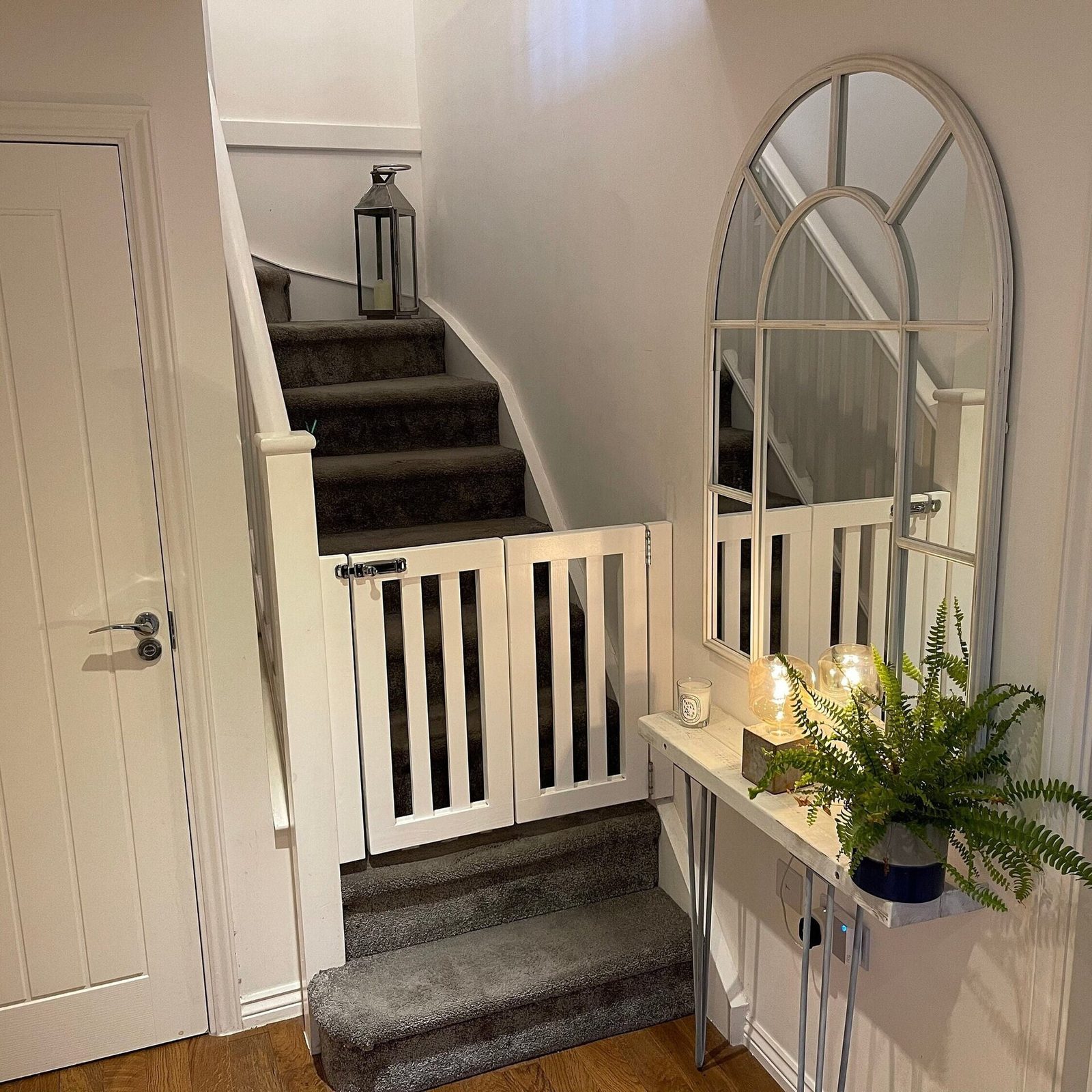 A white wooden double stair gate with a metal latch fitted across a grey carpeted staircase in a hallway, beside a white door and an arched mirror