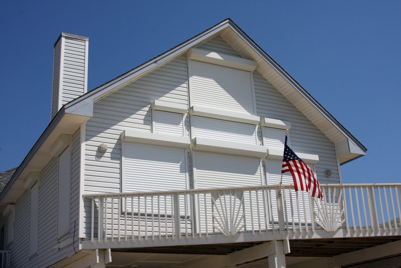 A pale grey beach house with roll-down storm shutters closed over its windows and doors, a white balcony railing and an American flag against a blue sky