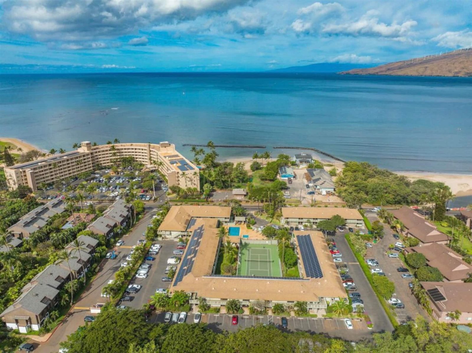 Aerial view of Kihei Bay Surf in Kīhei, a low-rise complex with solar panels on its roofs set around a tennis court and a pool, across a road from the beach, with a long rock wall in the shallow water and a dry ridge across the bay