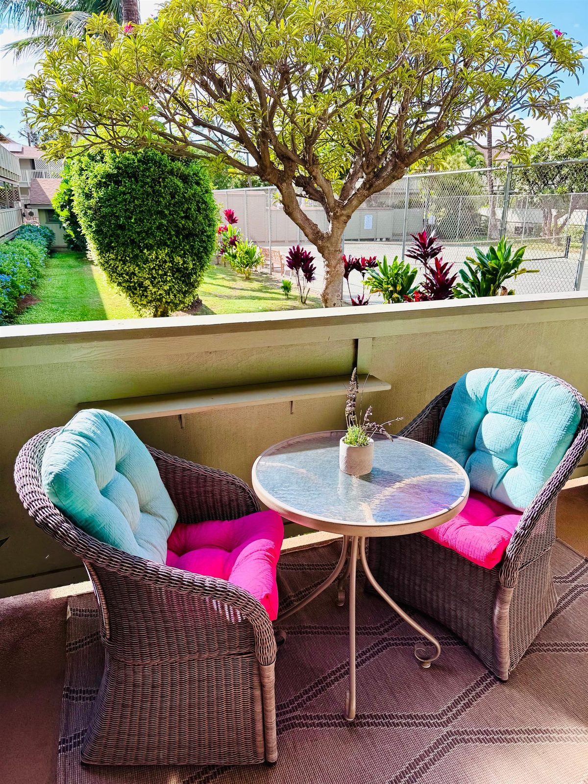 The studio’s lanai at Kihei Bay Surf, with two wicker chairs on turquoise and pink cushions and a round glass table, looking over a lawn, a tree and red-leaved plants to a fenced tennis court