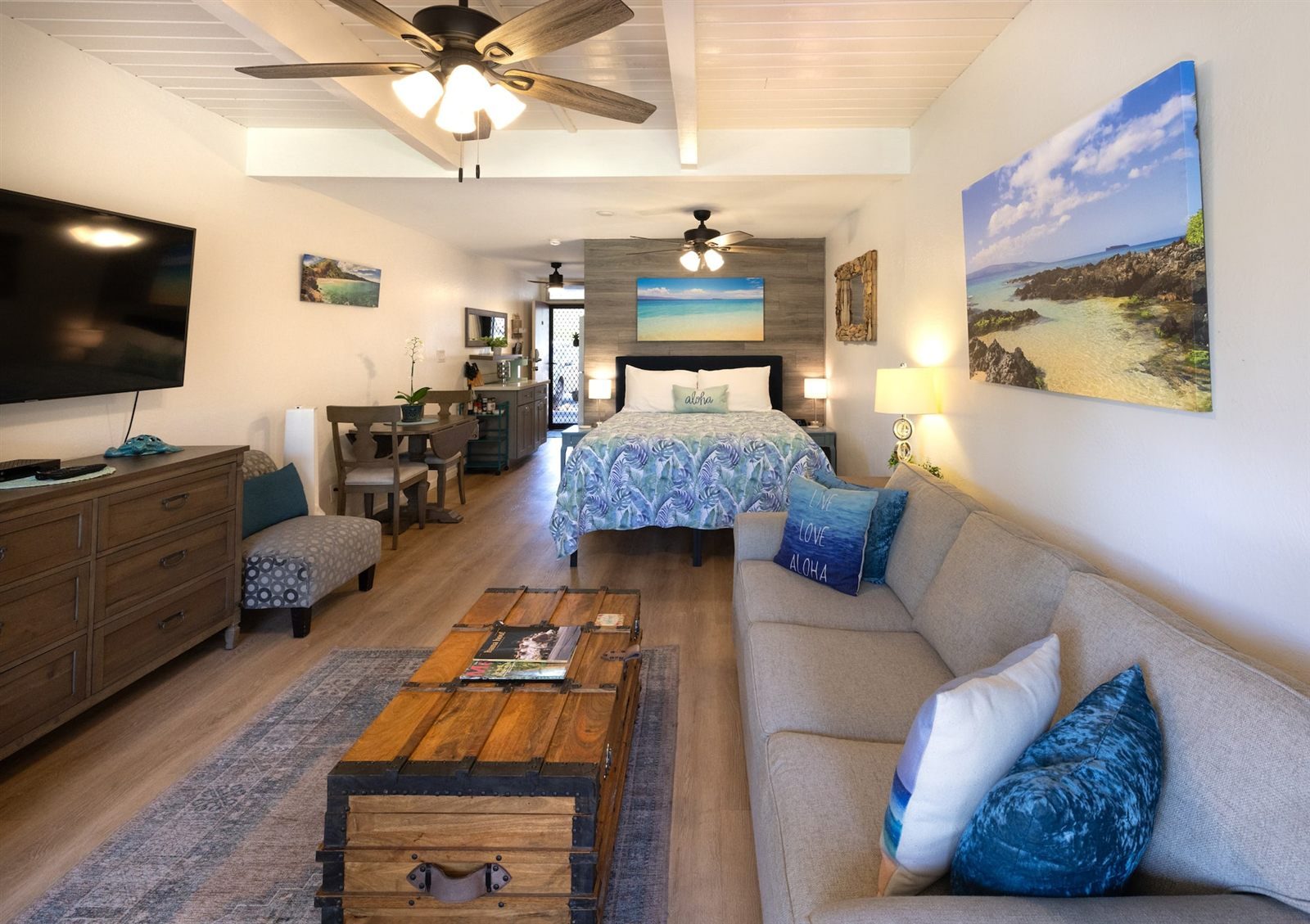The Couples Getaway studio, a long room with a grey sofa, a wooden trunk used as a coffee table, a dresser with a large television, a queen bed against a grey plank wall, ceiling fans and a small dining table near the kitchen