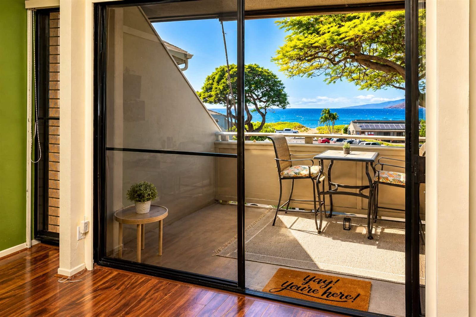 The sliding door from the Family Favorite onto its lanai, set with a high table and two tall chairs, looking over trees and a car park to the sea and the West Maui Mountains