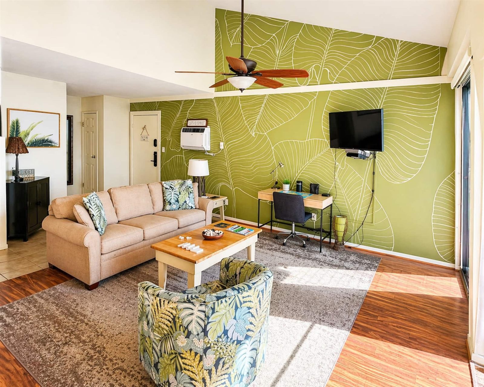 The living room of the Family Favorite, with a green wall patterned with large white palm-leaf outlines, a beige sofa, a leaf-print swivel chair, a wooden coffee table, a desk and a wall-mounted television under a ceiling fan