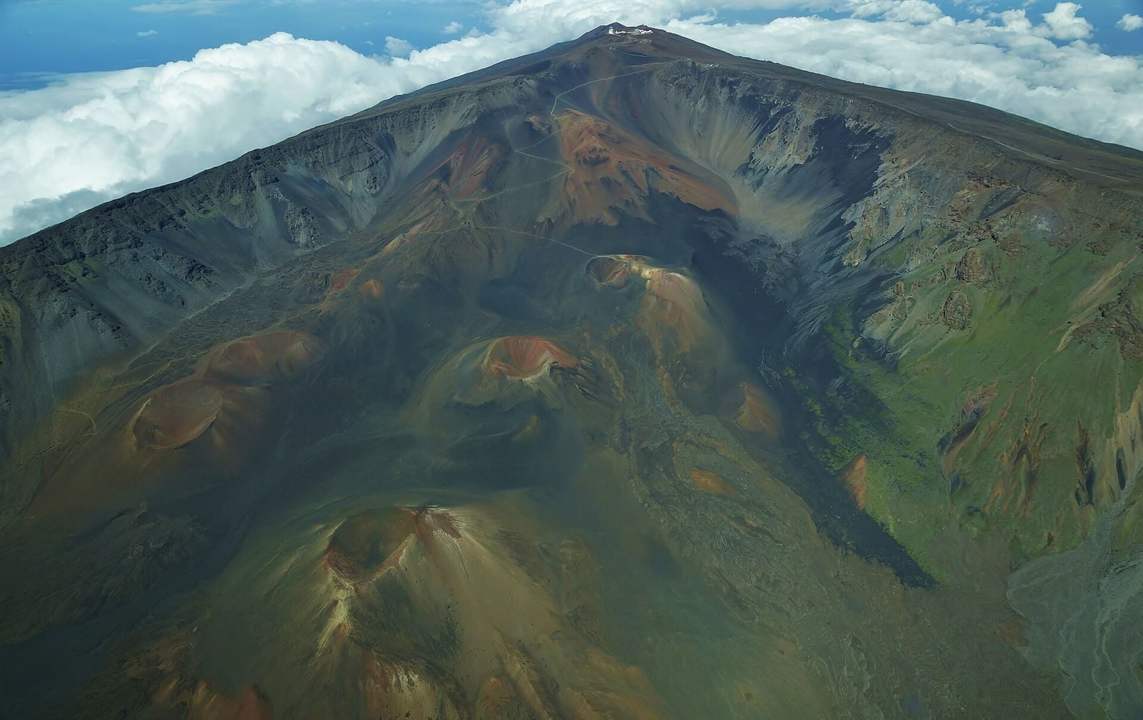 Aerial view of Haleakalā’s summit crater, a wide basin of red, brown and grey cinder cones below a steep summit ridge, with white observatory buildings on the peak and cloud beyond