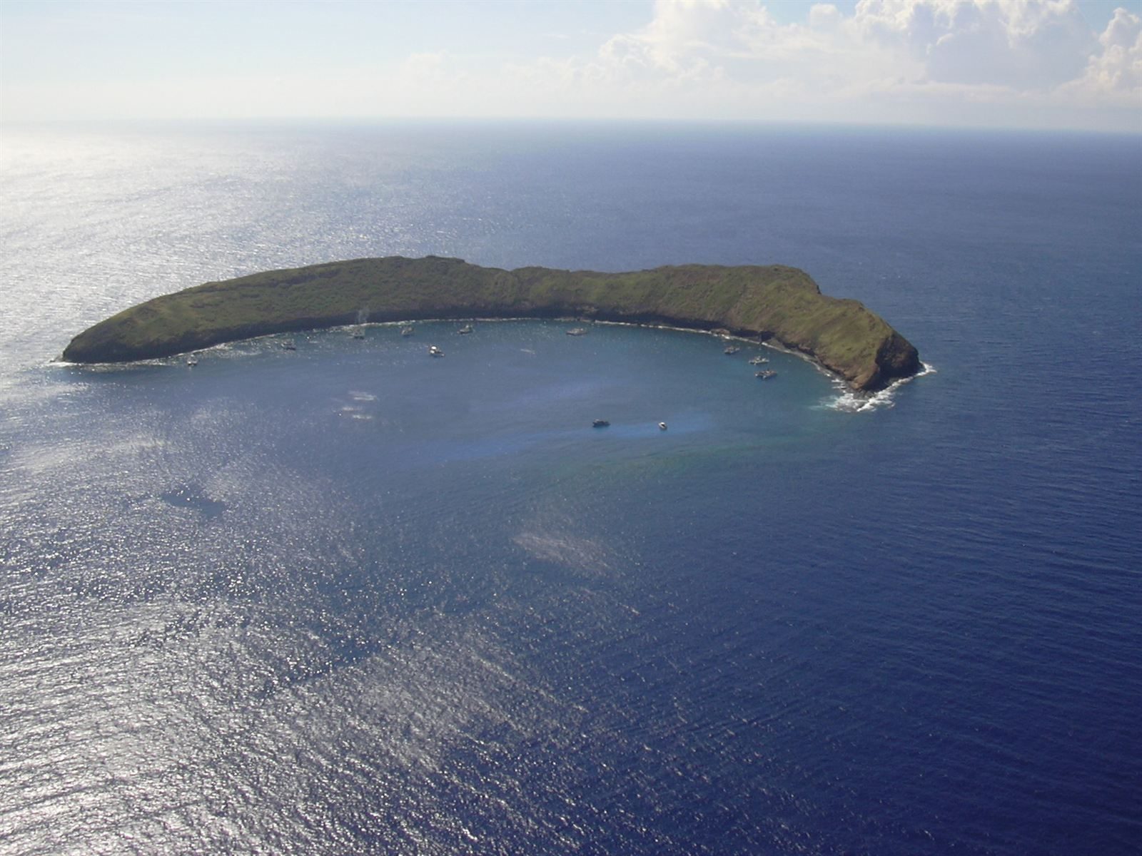 Molokini from the air, a crescent-shaped green islet in deep blue sea, with small boats moored in the calm water inside the curve of its crater rim