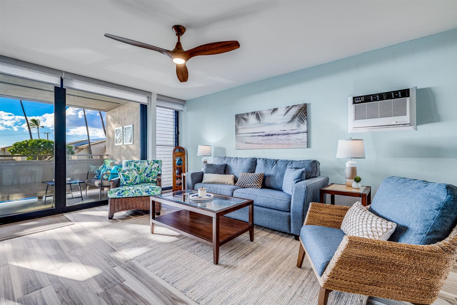 The living room of the Modern Ocean View, with pale blue walls, a blue sofa, two woven armchairs, a glass coffee table, a wall air-conditioner and a ceiling fan, and sliding doors to a lanai with palm trees beyond