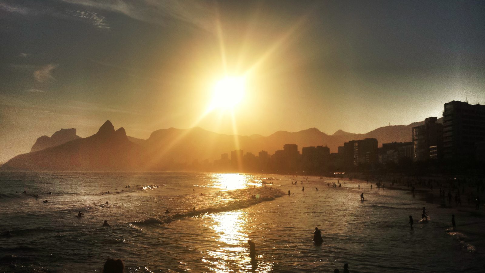 Sunset over Ipanema seen from the rocks at Arpoador, with the twin peaks of Dois Irmãos on the left, apartment blocks along the shore on the right and swimmers in the sea