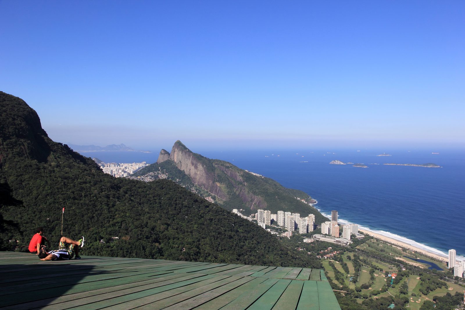 The wooden launch ramp on Pedra Bonita, with two people sitting on it, looking down over São Conrado's apartment towers, golf course and beach to the Dois Irmãos ridge and the Atlantic with its islands