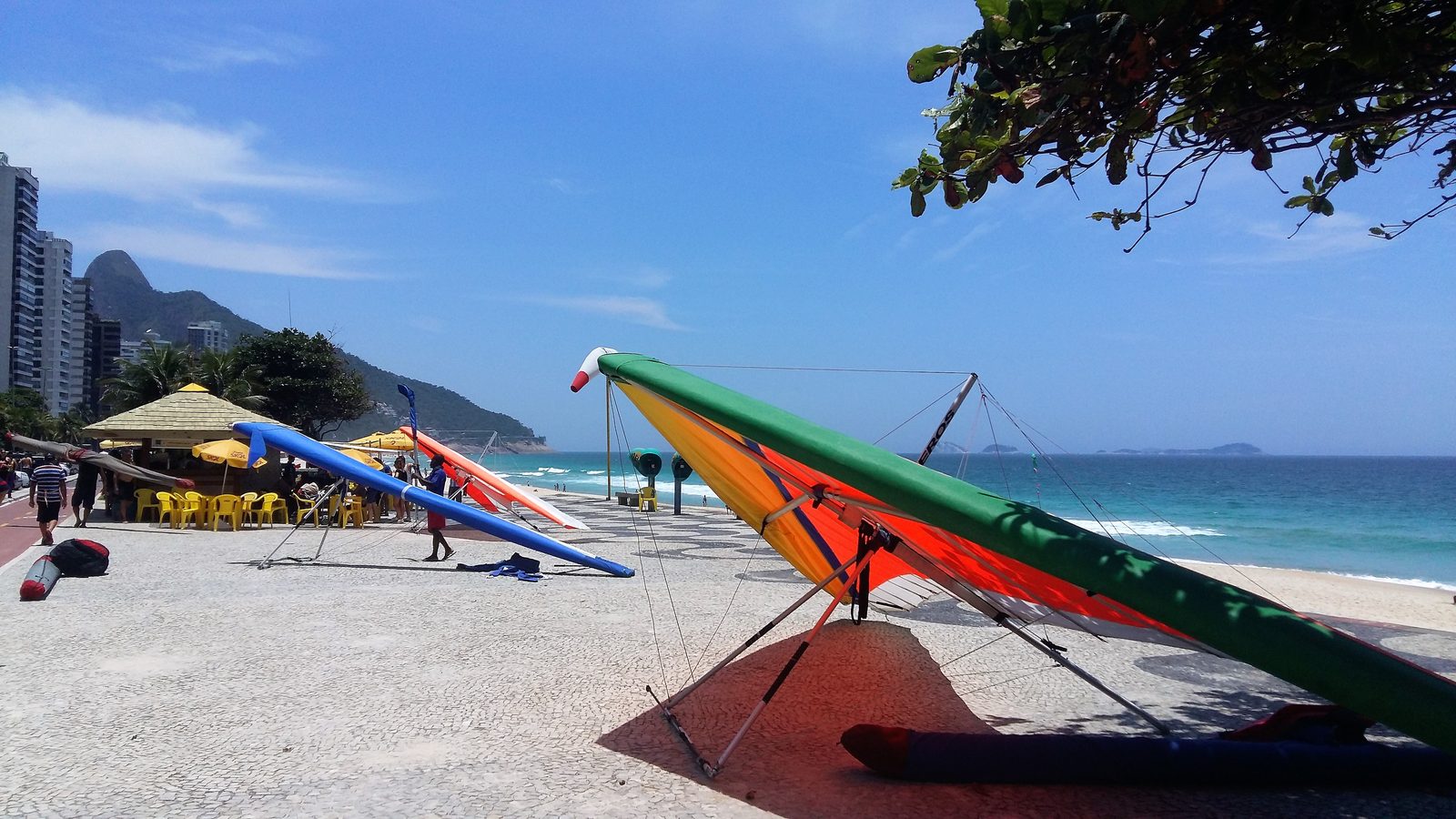 Hang-gliders resting on the patterned promenade beside São Conrado beach, near a thatched kiosk with yellow chairs, with apartment towers and a green hill behind