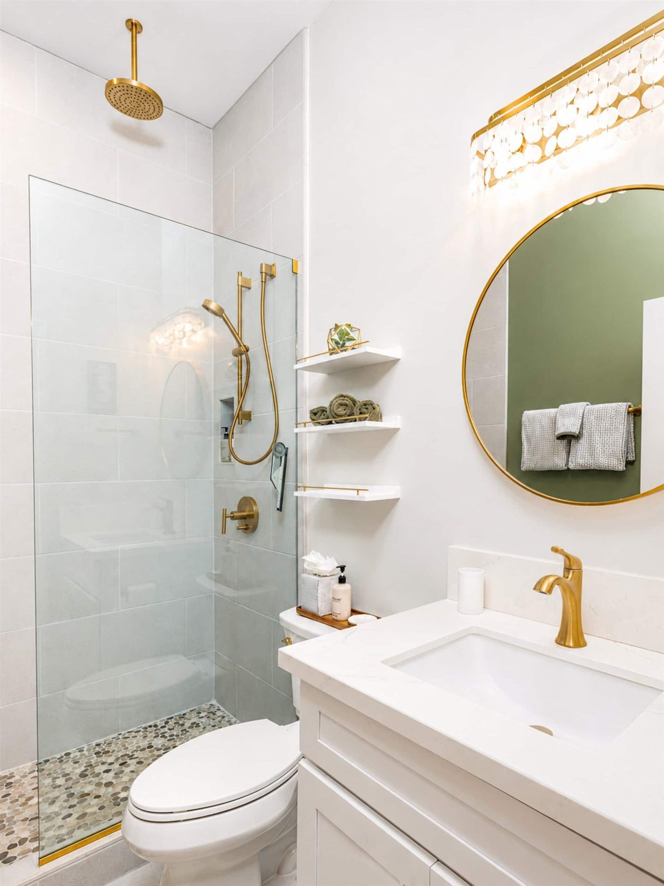 Bathroom with a glass-walled walk-in shower, a brass rain showerhead and fittings, a pebble-stone shower floor, a white vanity with a brass tap, a round brass-framed mirror and a shell-disc light.