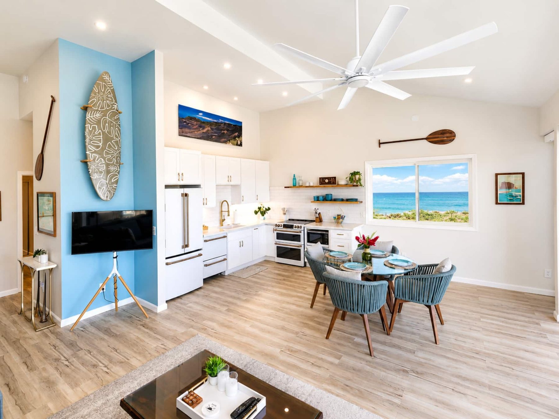 Open-plan room with a white kitchen along the back wall, a round glass dining table with four blue woven chairs, a blue accent wall holding a decorated surfboard, a large white ceiling fan and a window facing the ocean.
