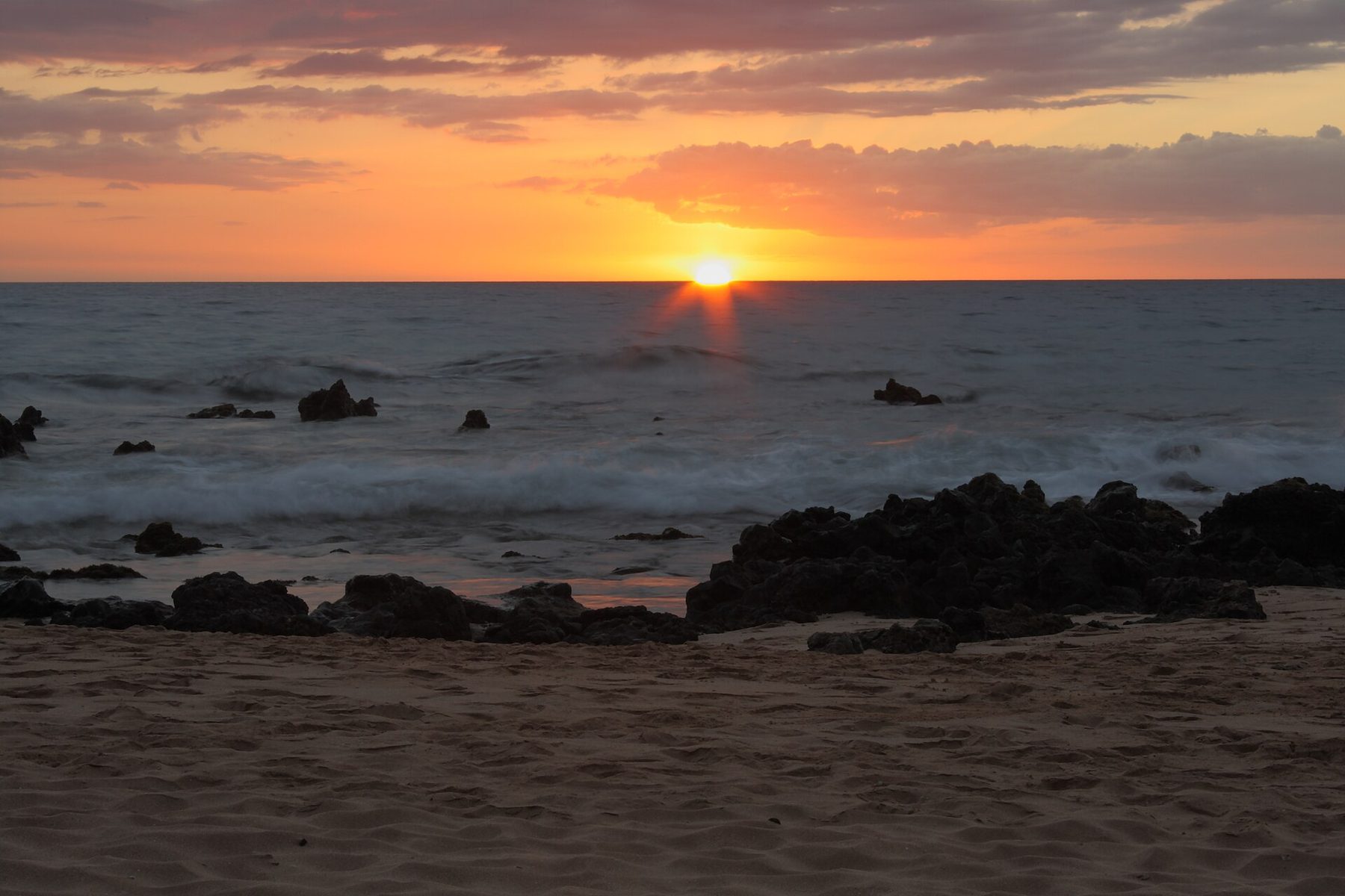 Sun touching the ocean horizon under orange and grey clouds, with dark lava rocks, blurred surf and rippled sand in the foreground.