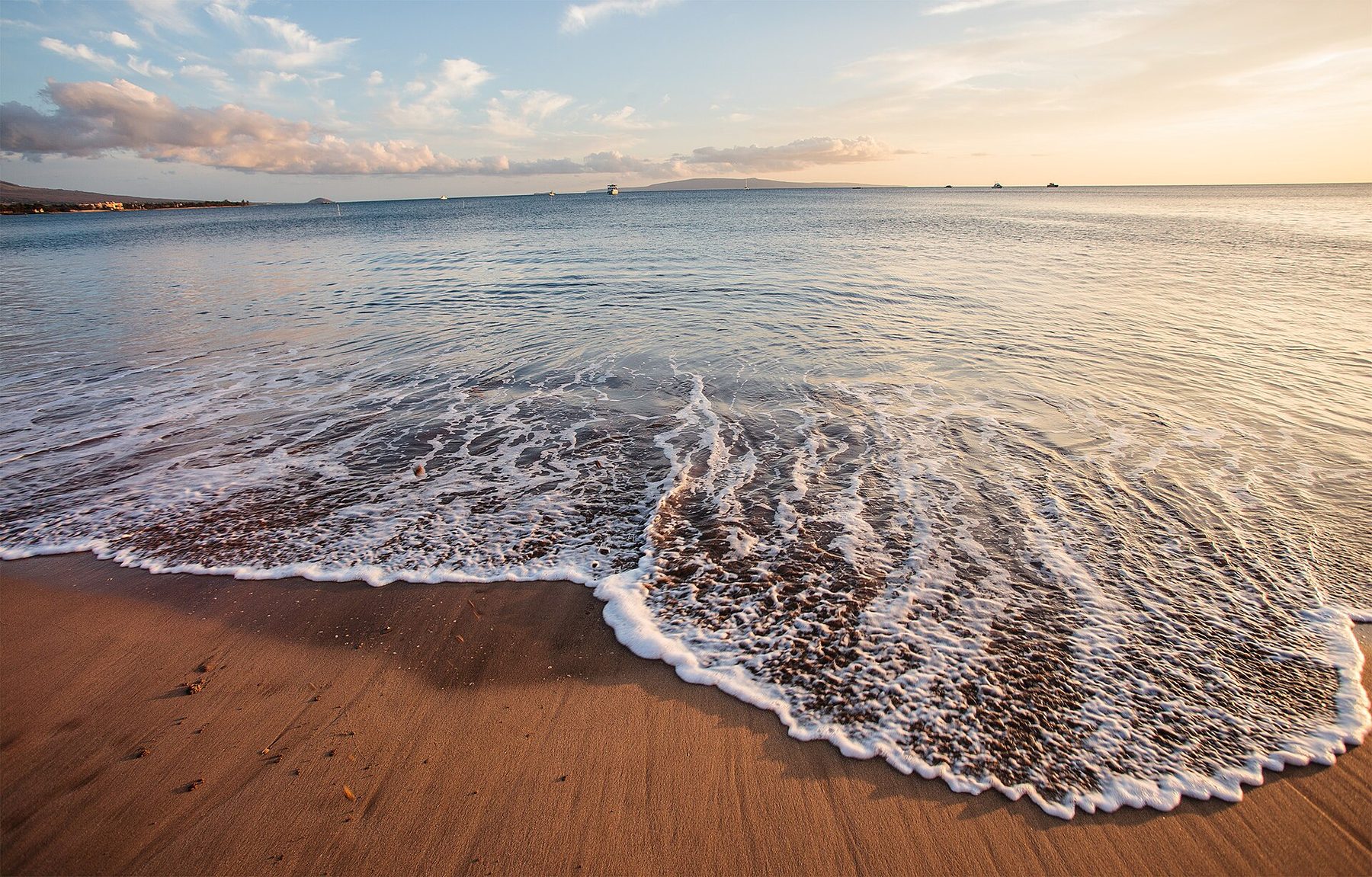 Foamy edge of a small wave spreading over wet brown sand at sunset, with calm water, several boats, a low island on the horizon and pink-lit clouds.